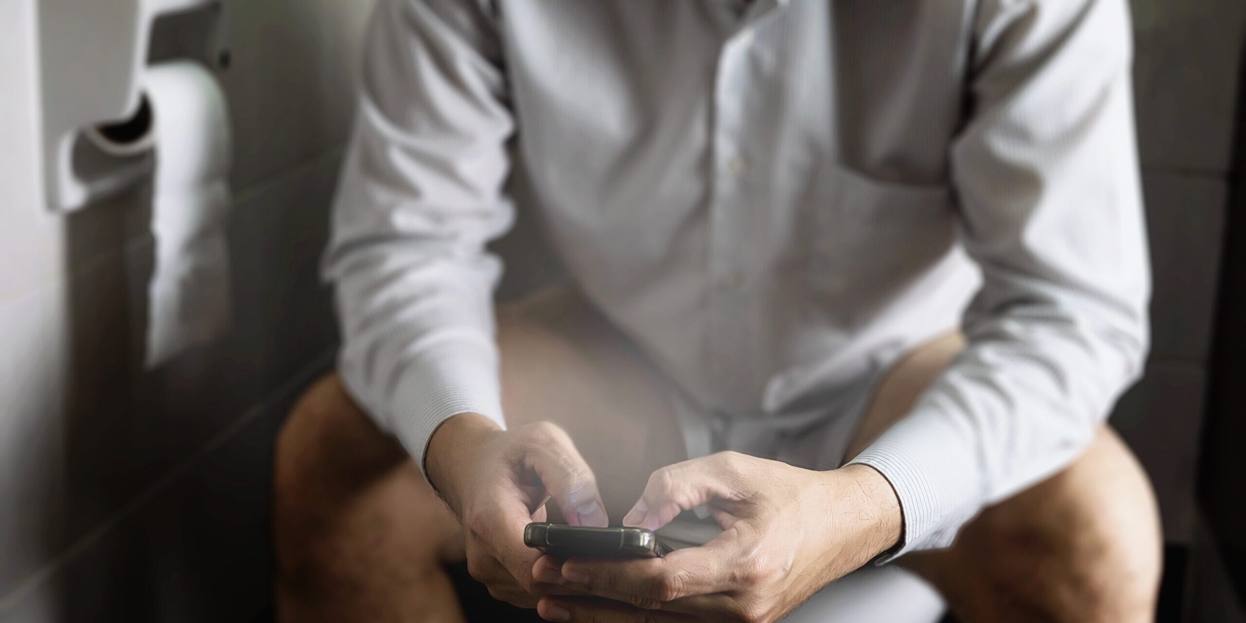 Man sitting on toilet bowl while playing mobile phone - health problem concept